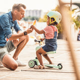 A parent walking beside a toddler in a helmet riding a sit-on scooter in the park